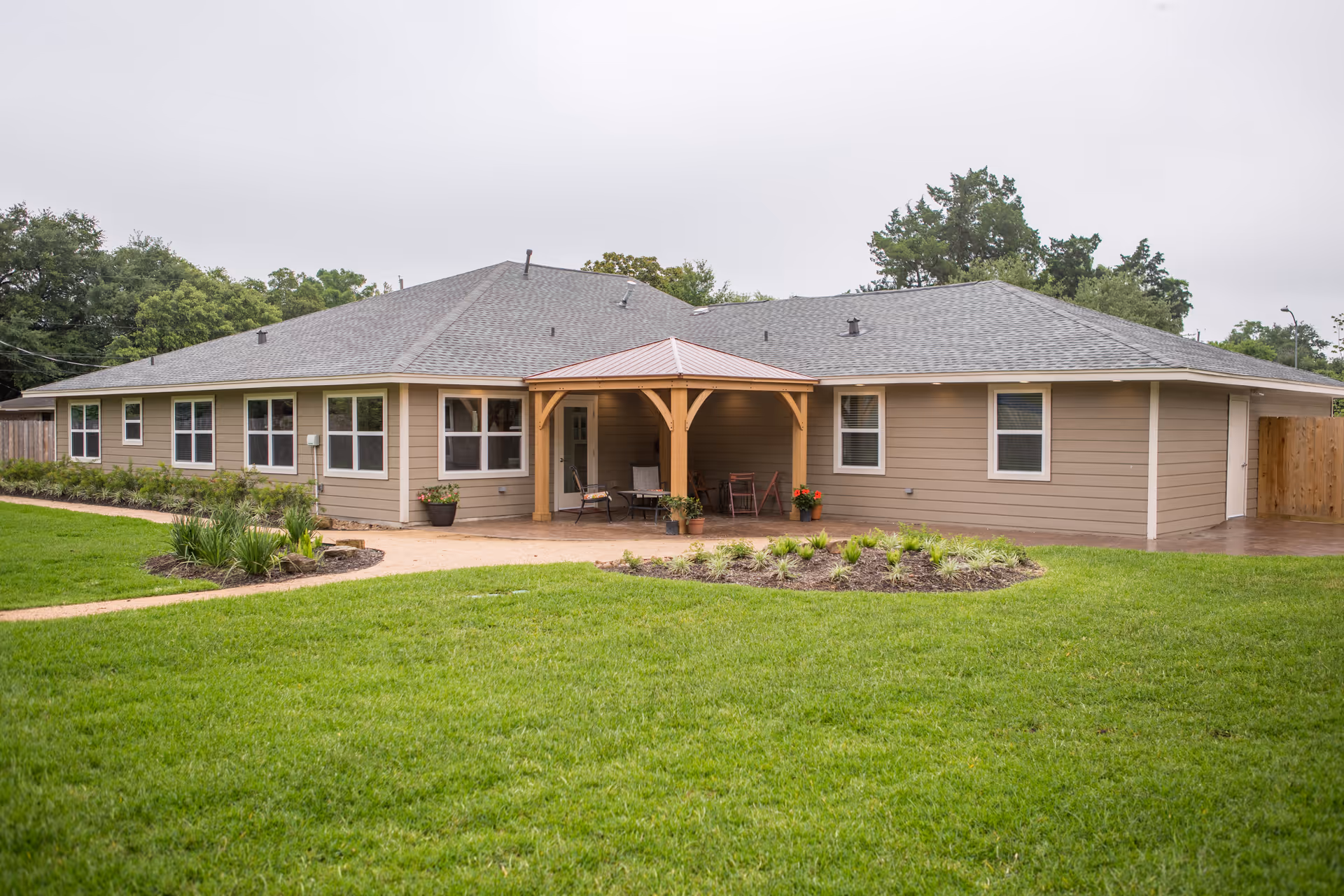 Exterior view of a single-story assisted living facility building with beige siding and a gray shingled roof. The building features multiple windows and a covered patio area with wooden posts and outdoor seating. The surrounding area includes a well-maintained green lawn and landscaped garden beds.