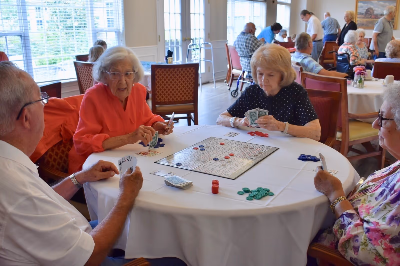 Four elderly people sitting around a round table playing a board game with cards and colored chips in a well-lit room with large windows and other people in the background.