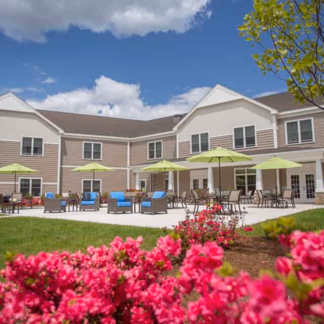 Outdoor patio area of a senior living facility with beige two-story building in the background, green umbrellas shading tables and chairs, blue cushioned seating, and vibrant pink flowers in the foreground under a partly cloudy blue sky.