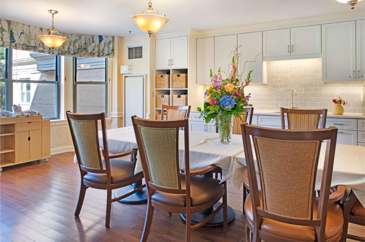 Bright dining room with a table and wooden chairs, a floral centerpiece, large windows and a white kitchen area in the background.