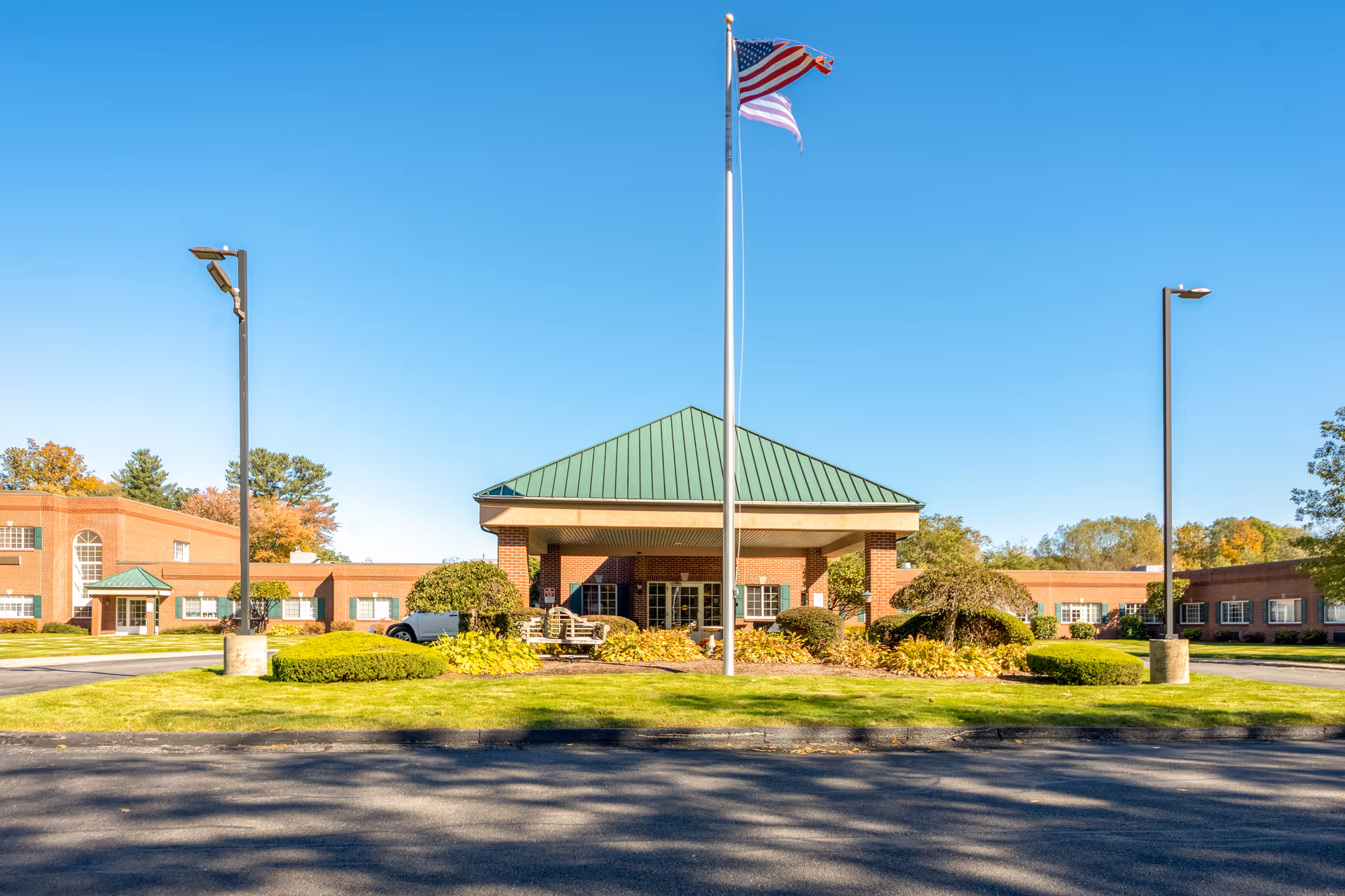 Front exterior view of a senior living facility with a green metal roof entrance, an American flag on a flagpole in the center, surrounded by landscaped bushes and trees under a clear blue sky.