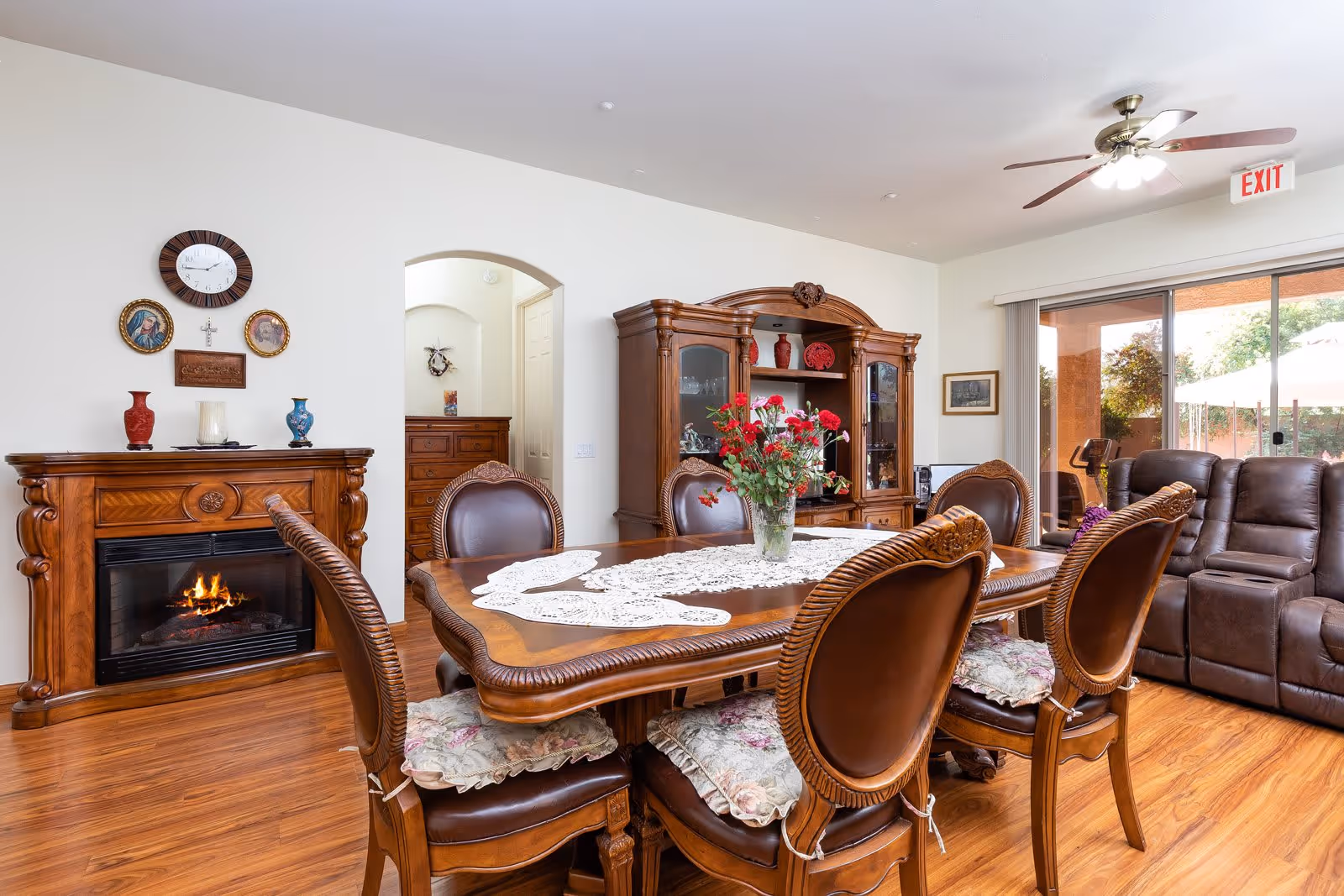 A cozy dining and living area featuring a wooden dining table with six cushioned chairs, a vase with red flowers as a centerpiece, a wooden fireplace with a small fire burning, a wooden cabinet with decorative items, a ceiling fan with lights, and a sliding glass door leading to an outdoor patio with an umbrella and exercise equipment.