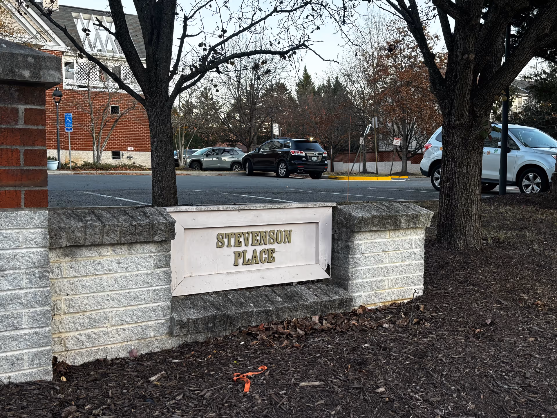 A low stone sign reading 'STEVENSON PLACE' sits in landscaping in front of a parking lot with trees and parked cars.