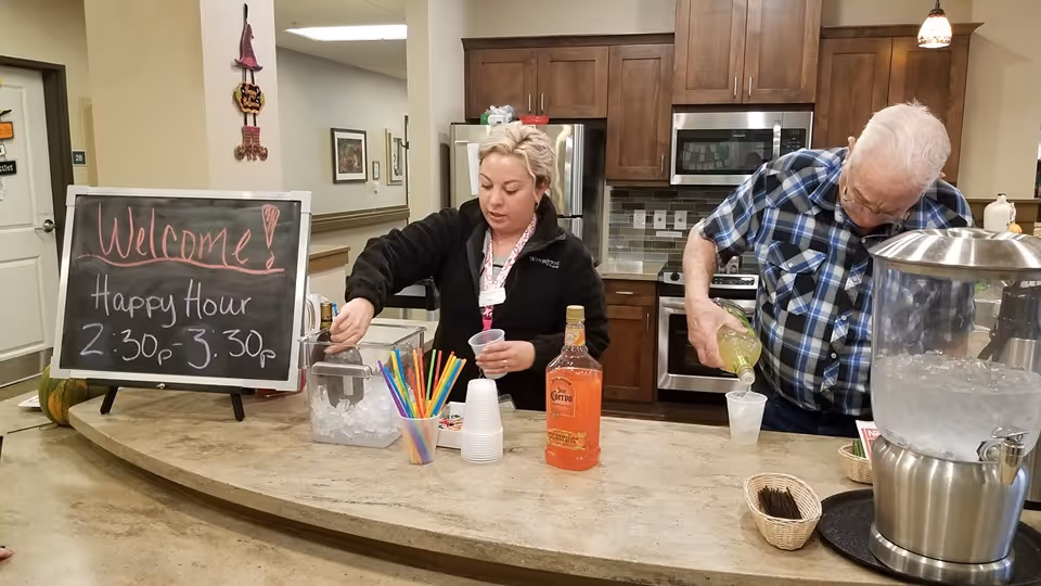 A woman and an elderly man are preparing drinks behind a counter in a kitchen area. The woman is scooping ice into a cup while the man is pouring a beverage from a bottle. On the counter, there are colorful straws, plastic cups, a bottle of orange liquor, and a large water dispenser. A chalkboard sign on the left side reads 'Welcome! Happy Hour 2:30p - 3:30p'. The background shows wooden kitchen cabinets, a microwave, and a refrigerator.