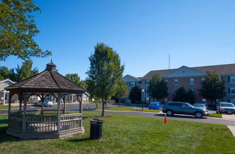 Outdoor scene at Gateway Village featuring a wooden gazebo on a grassy area with a trash can nearby. In the background, there are several trees, a parking lot with multiple cars, and a multi-story residential building under a clear blue sky.
