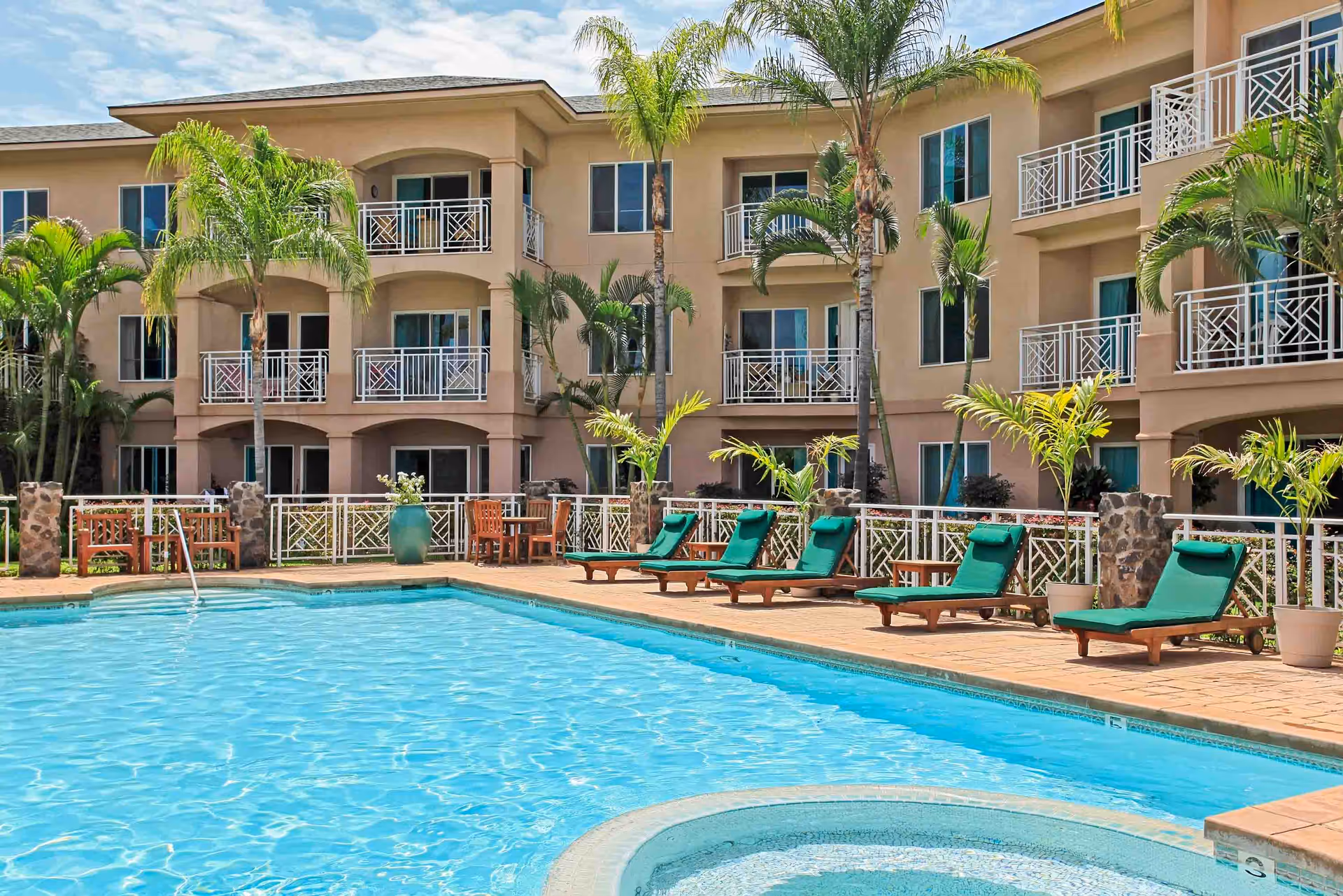 Resort-style outdoor swimming pool with green lounge chairs, palm trees, and a three-story residential building with balconies.