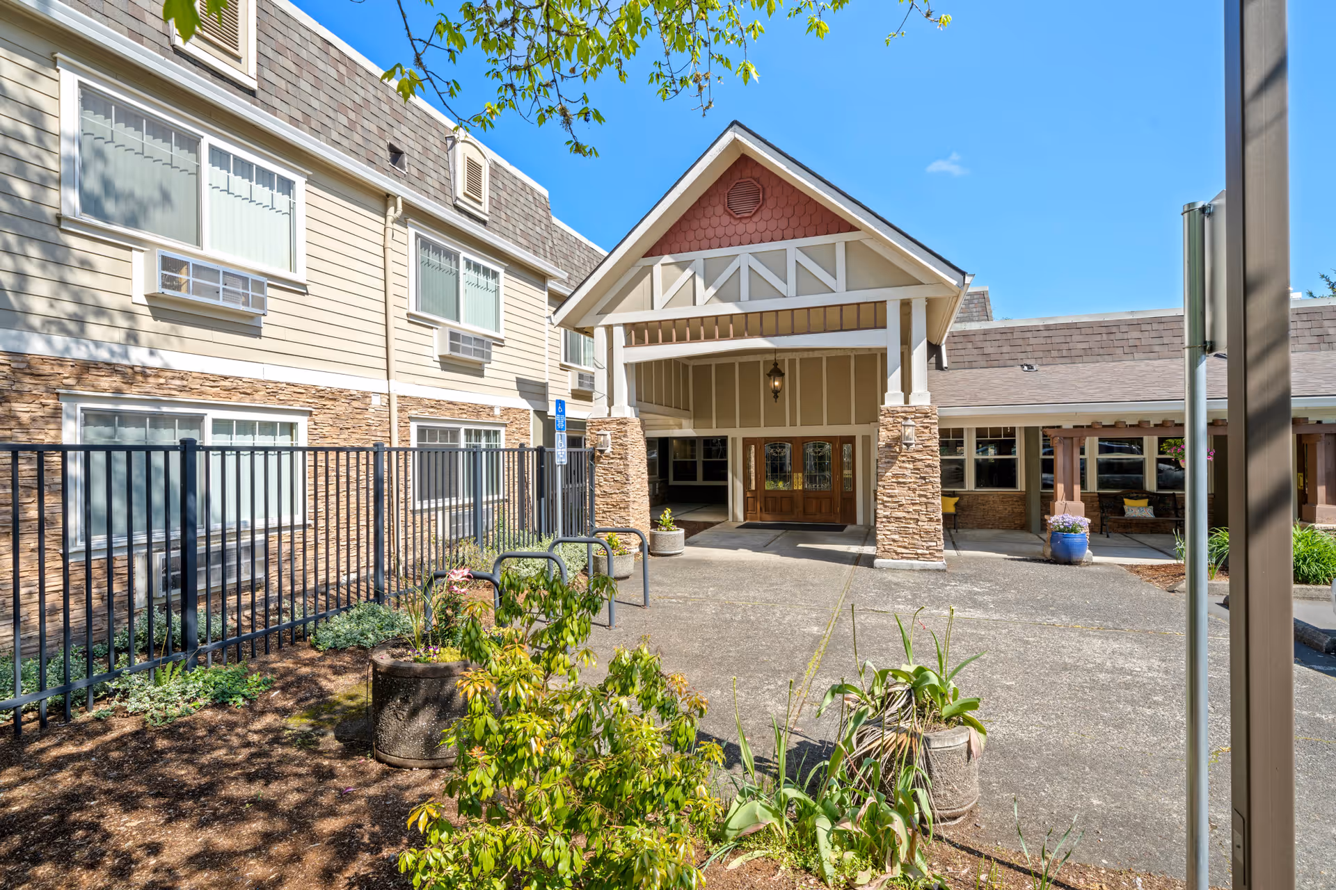 Exterior view of the entrance to a senior living facility named Royalton Place, featuring a covered entryway with stone pillars, large windows, and surrounding landscaping with plants and trees under a clear blue sky.