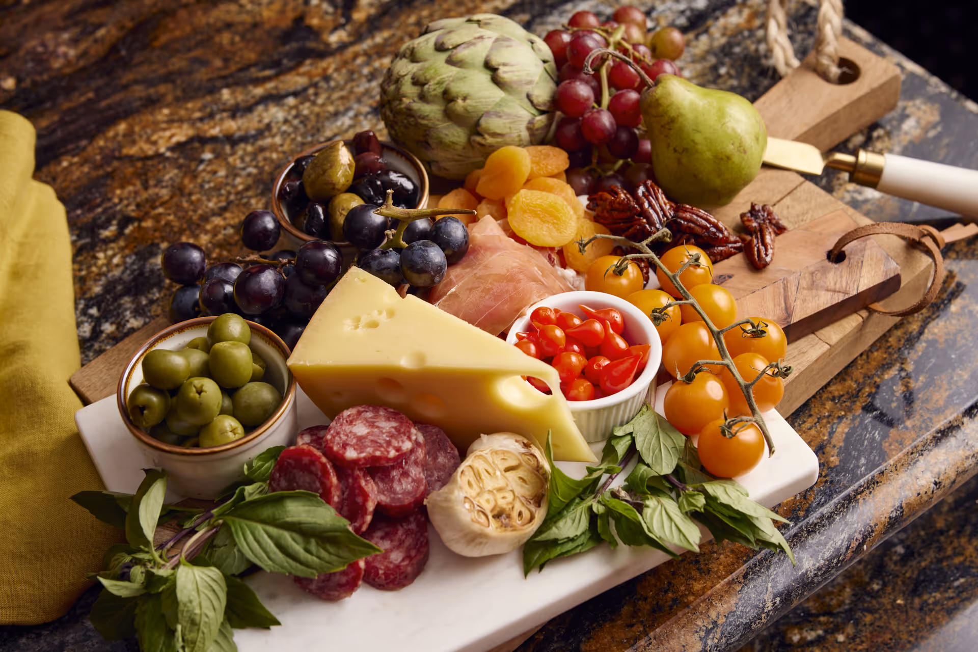 A charcuterie board on a marble surface featuring a wedge of Swiss cheese, slices of salami, green and black olives, dried apricots, cherry tomatoes on the vine, pecans, a pear, an artichoke, a head of garlic, fresh basil leaves, and a small bowl of red pickled peppers.
