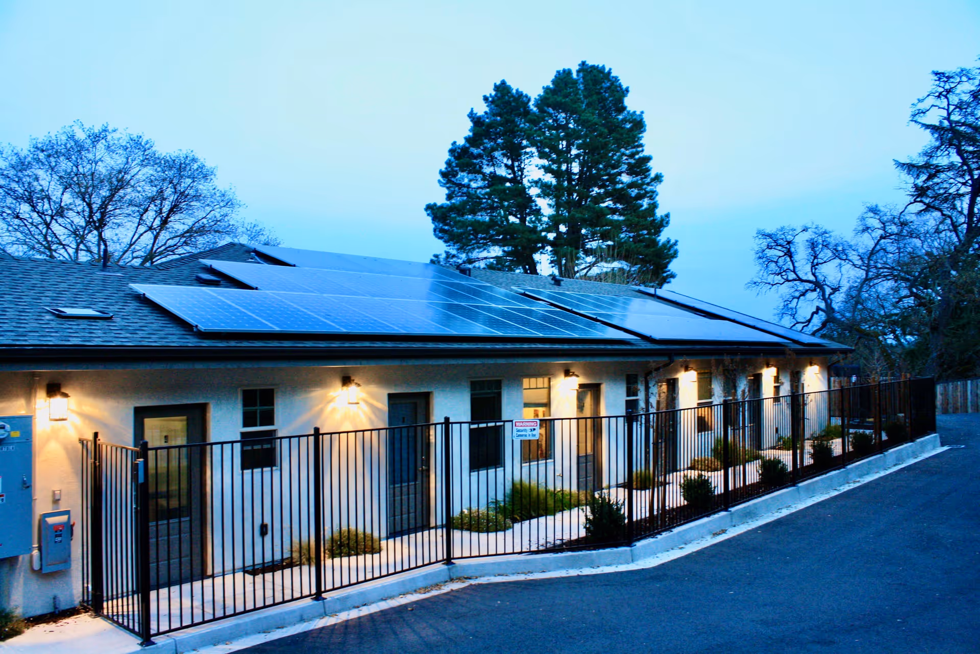 Row of ground-level assisted living units with solar panels on the roof, exterior lights, and a metal fence along the sidewalk.