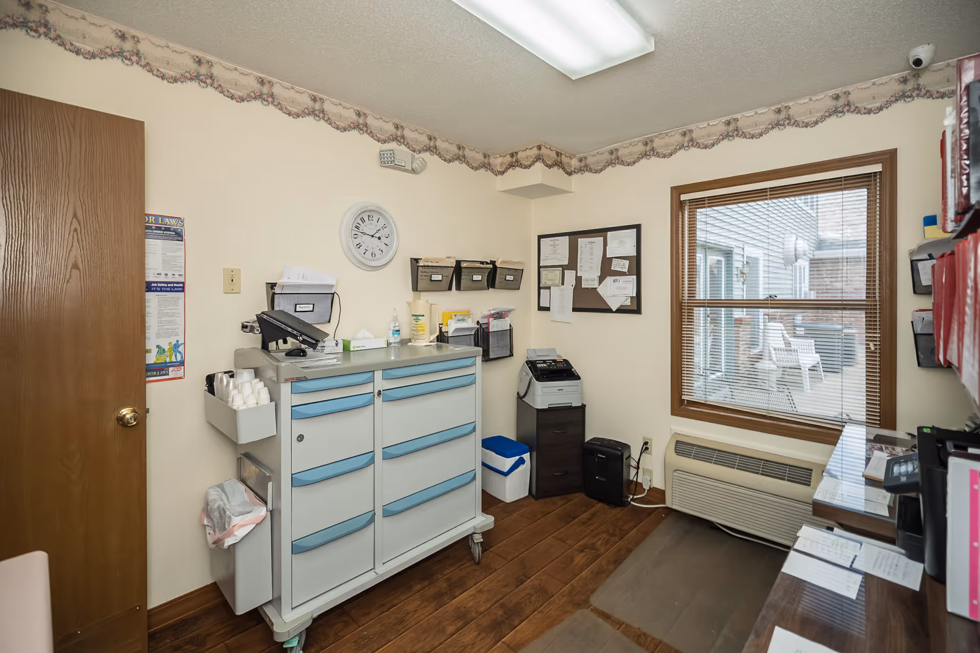 Small office room with a wooden door, a white and blue medical cart with supplies, a clock on the wall, a bulletin board with papers, a printer on a small cabinet, a window with blinds showing outdoor chairs, and a desk with organized files and papers.