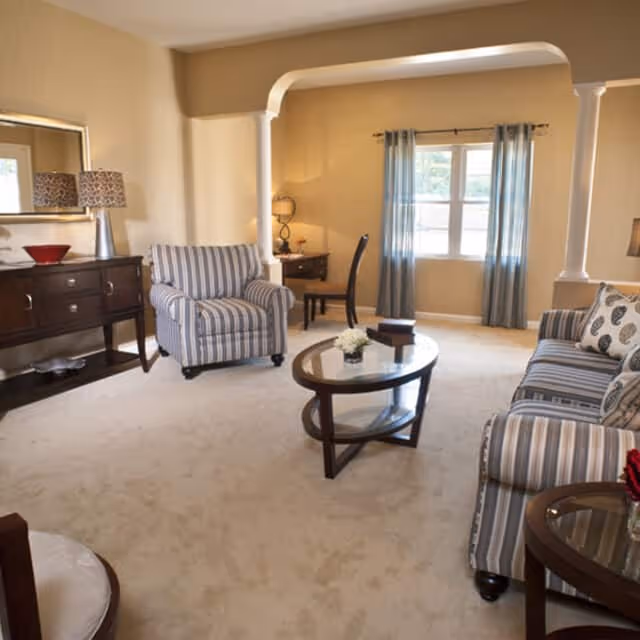 Bright furnished living room with a striped sofa and armchair, oval glass coffee table, sideboard, and a window with curtains.