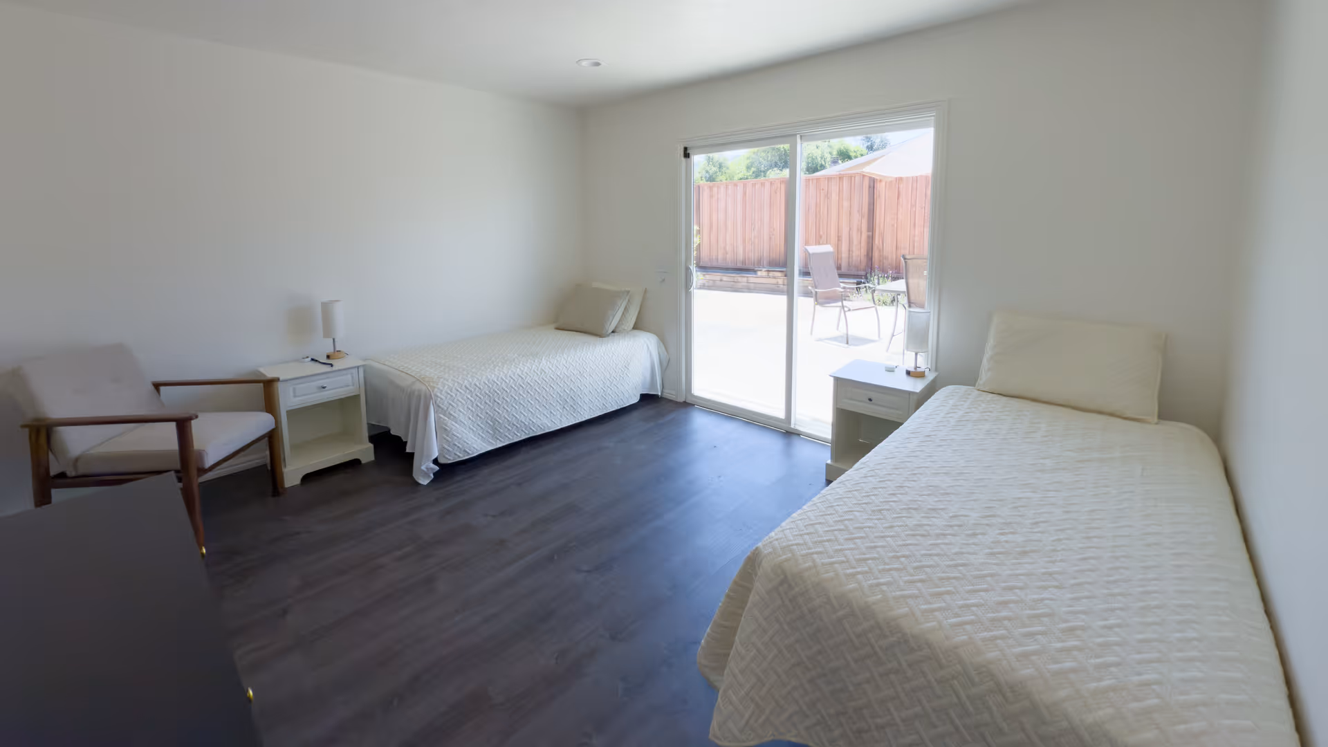 Bright, minimal bedroom with two twin beds, nightstands, a chair, and a sliding glass door opening to a fenced patio.