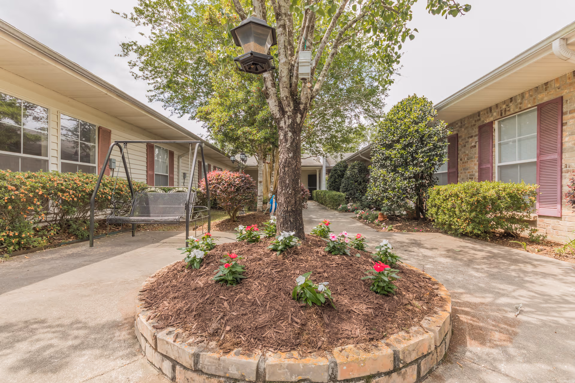 Courtyard with a central tree planter and flowers, a swinging bench, walkways, and surrounding assisted-living building.