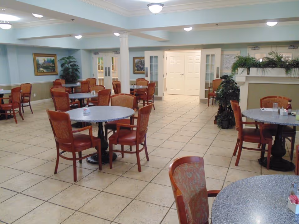 Spacious dining area with round tables and upholstered chairs arranged on a tiled floor inside a senior living facility.