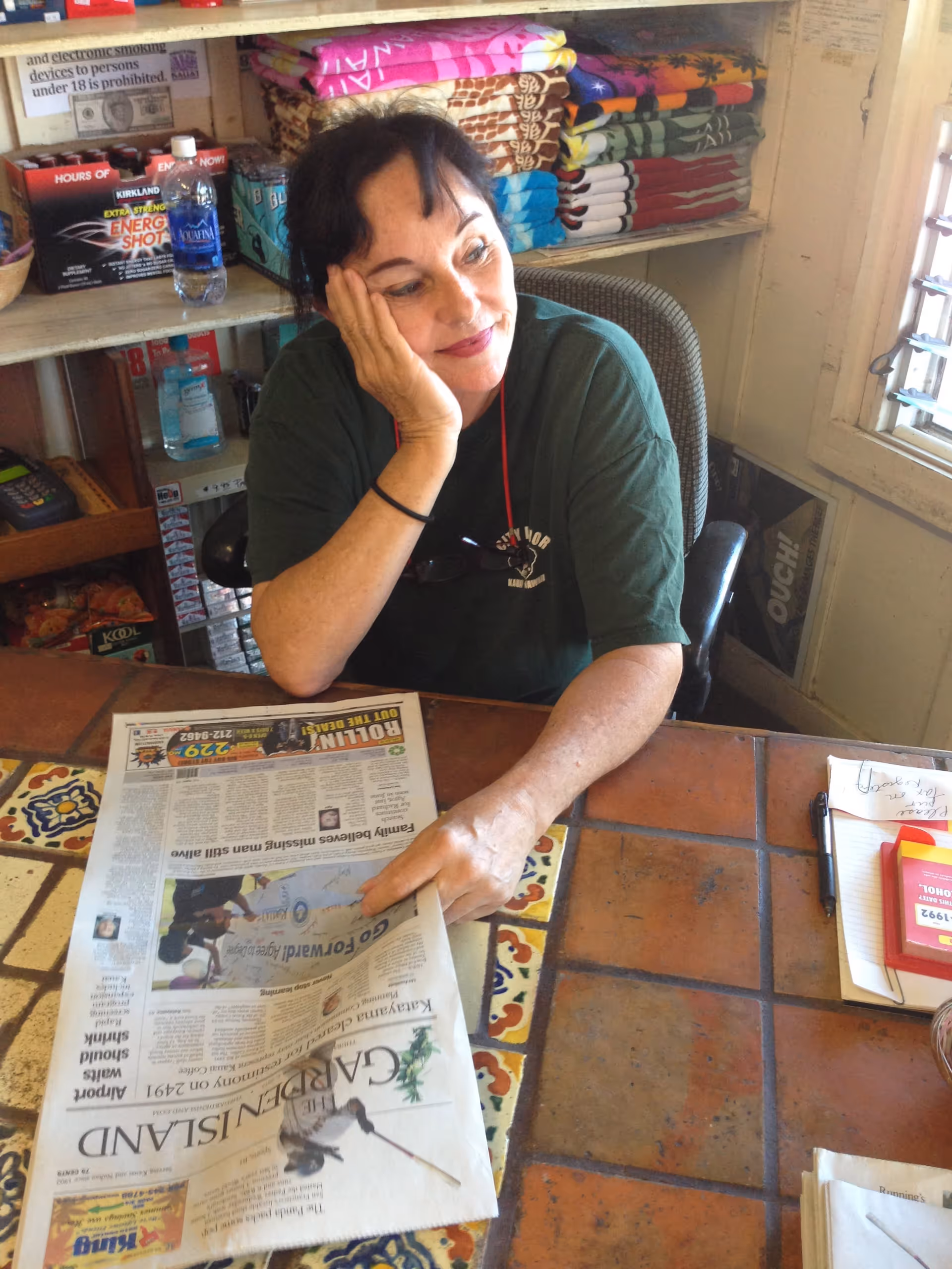 A woman with dark hair wearing a green shirt sits at a tiled table, resting her head on one hand and holding a newspaper with the other. Behind her are shelves with folded colorful towels and various items including a water bottle and energy shots. The setting appears to be a cozy indoor space with a window to the right.