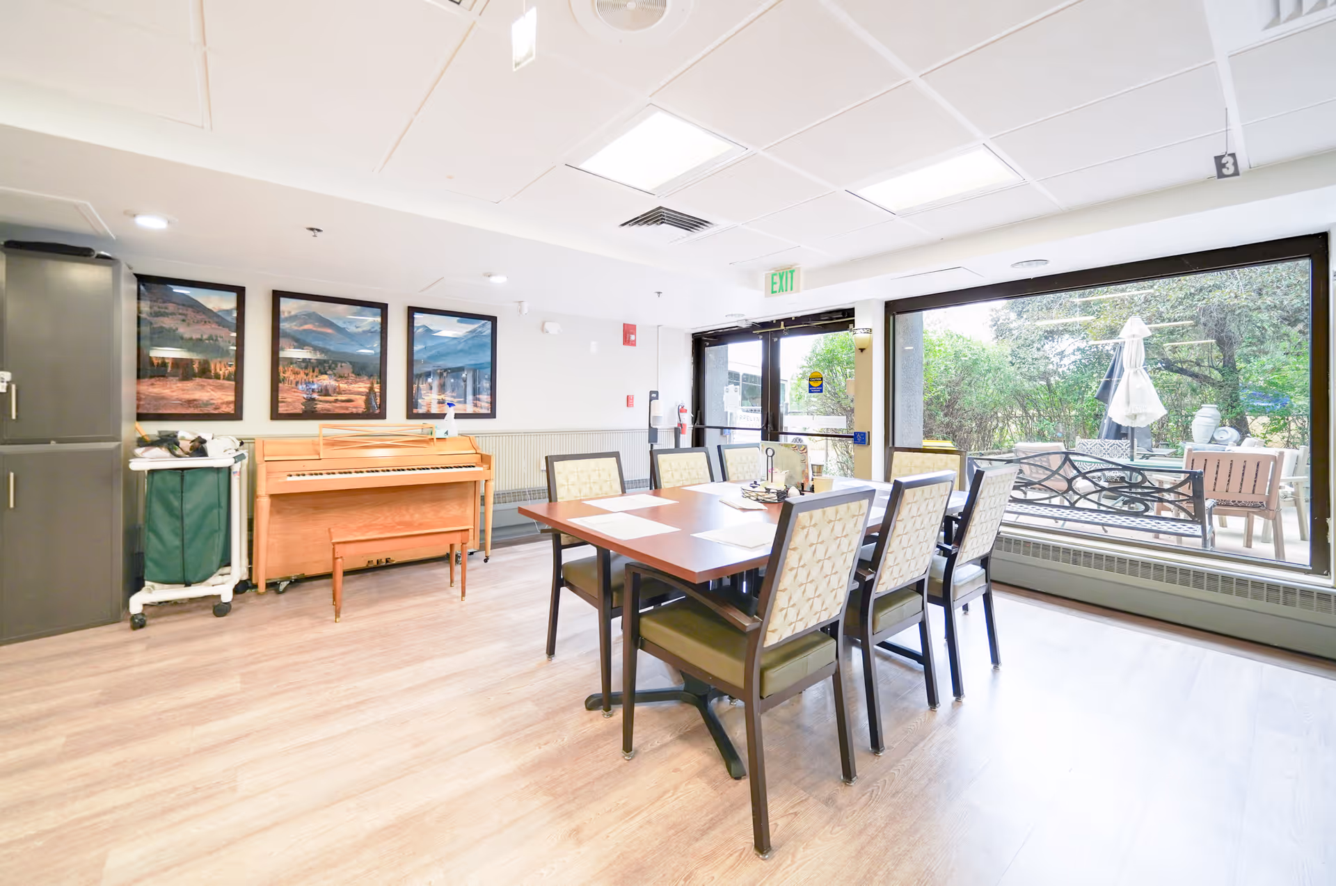 A bright room with a rectangular wooden table surrounded by six cushioned chairs. Behind the table is a wooden piano with a bench, and three framed landscape pictures hang on the wall above it. Large windows on one side provide a view of an outdoor patio area with benches, tables, and umbrellas. The room has light-colored wood flooring and a white ceiling with recessed lighting.