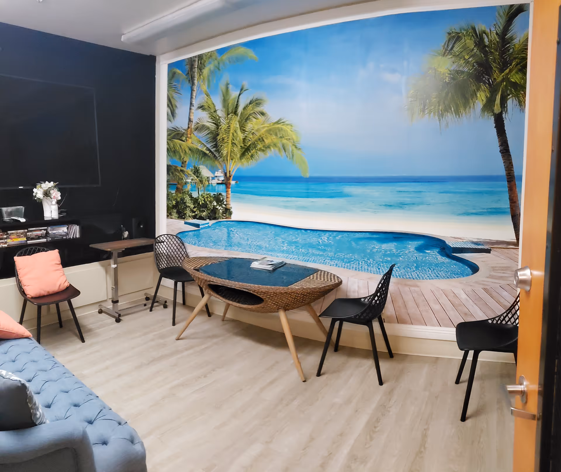 A cozy living room with a large wall mural depicting a tropical beach scene with palm trees and a pool. The room has a blue tufted sofa, a wicker coffee table with a glass top, three black chairs, a small side table with a vase of flowers, and a flat-screen TV mounted on a black wall.