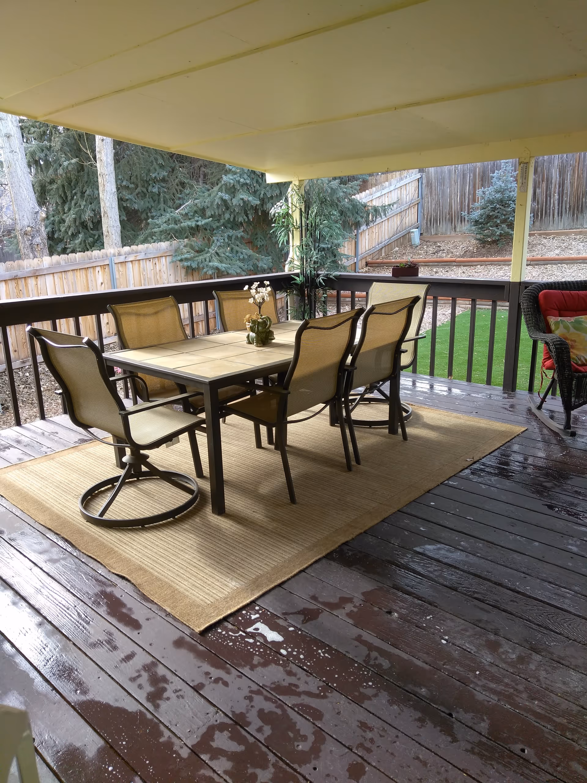 Covered outdoor wooden deck with a dining table and six chairs on a rug, overlooking a fenced backyard.