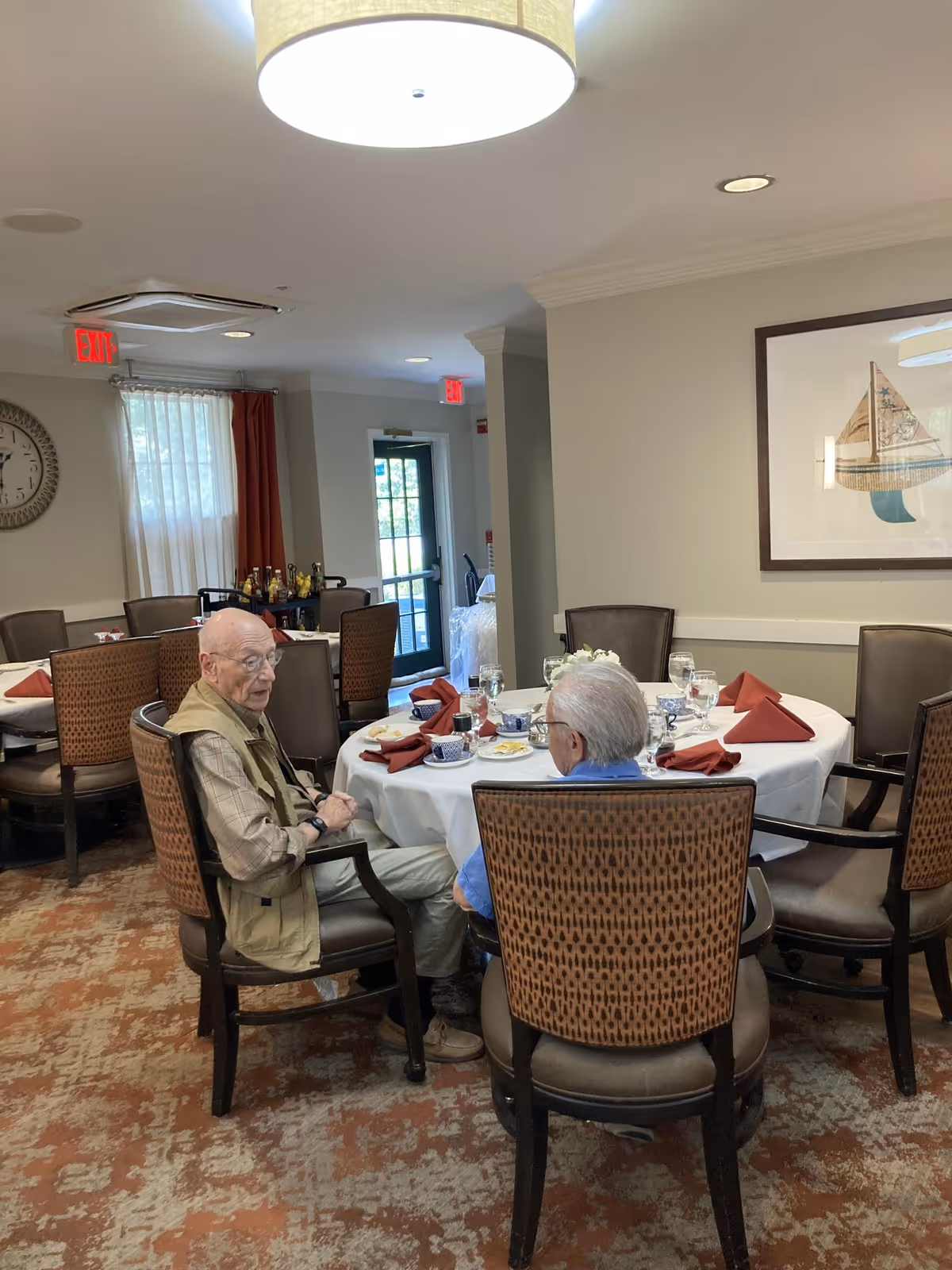Two elderly men sitting and talking at a round dining table set with white tablecloth, red napkins, plates, cups, and glasses in a well-lit dining room with patterned carpet, wall art, and exit doors in the background.