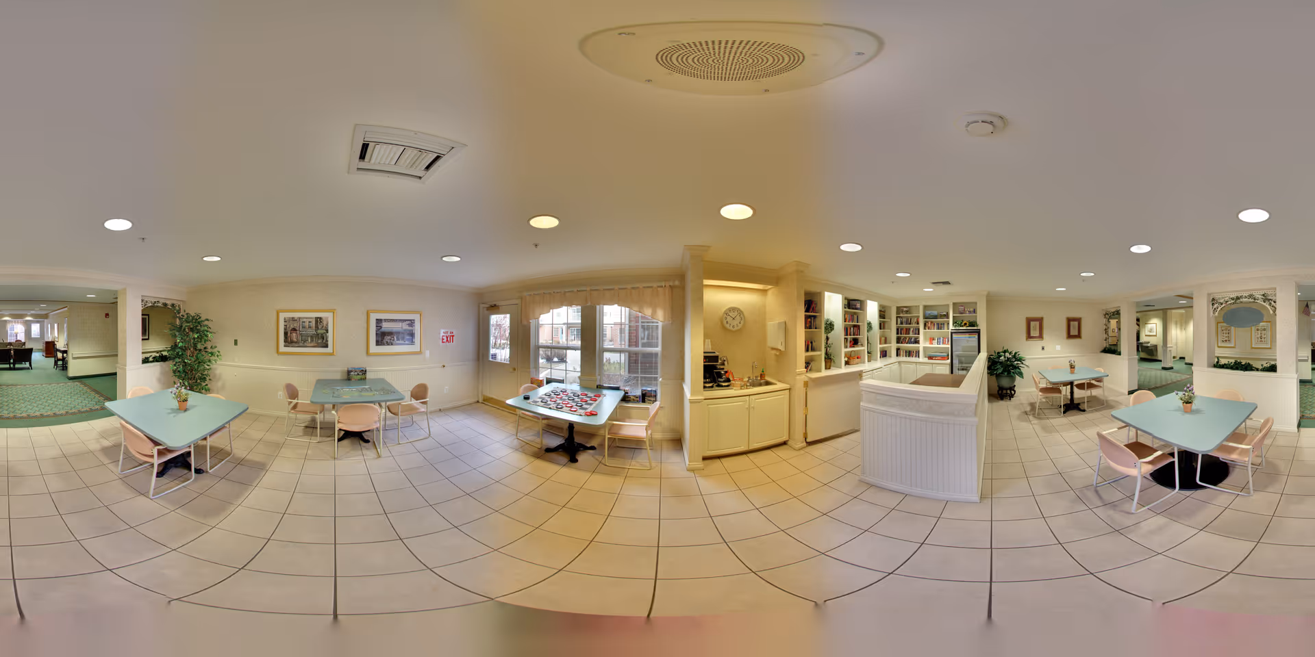 A spacious, well-lit common area in a senior living facility with multiple tables and chairs arranged for socializing or activities. The room features tiled flooring, large windows with sheer curtains, built-in shelving with books and games, and a small kitchenette area with a sink and coffee maker. The walls are decorated with framed pictures and there are plants placed around the room, creating a welcoming atmosphere.