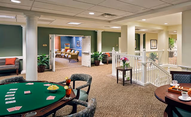 Interior view of a senior living facility common area with a card table set up with playing cards, snacks, and drinks. There are upholstered chairs around the table, a small round table with coffee cups and a tray, and a seating area with benches and potted plants. In the background, double doors open to another room with additional seating and framed artwork on the walls.