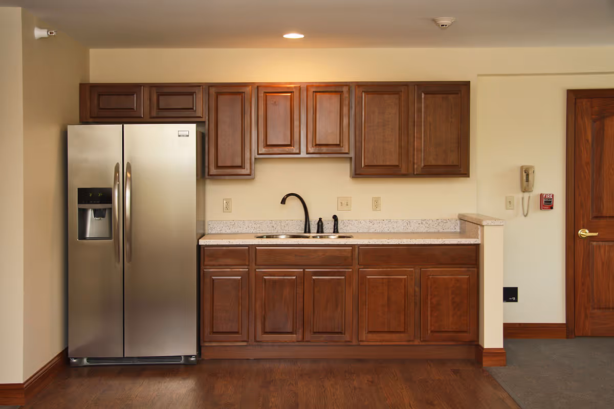 Small kitchen area with a stainless steel side-by-side refrigerator, dark wooden cabinets, a countertop and a sink with faucet.