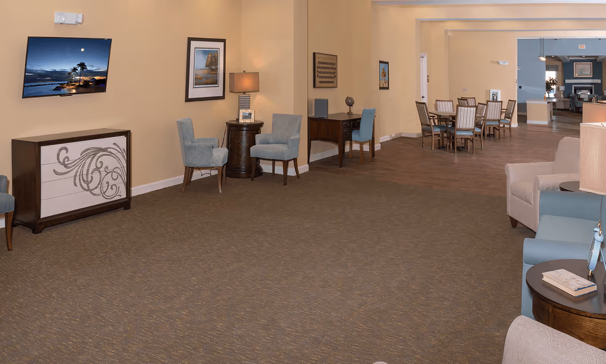 A spacious interior common area with beige walls and carpeted floor. The room features a wall-mounted TV displaying a scenic beach image, two light blue armchairs around a small round table with a lamp, a wooden desk with a blue chair and a globe, and a dining area with a table and chairs in the background. Additional seating with armchairs and side tables is visible in the foreground and background.