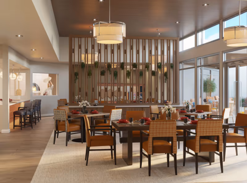 A modern dining room with multiple tables set with red plates and cups. The room features wooden chairs with orange cushions and patterned backs, a wooden slatted divider with hanging plants, large windows letting in natural light, and pendant lights hanging from the ceiling. There is a bar area with stools to the left and an outdoor seating area visible through glass doors on the right.
