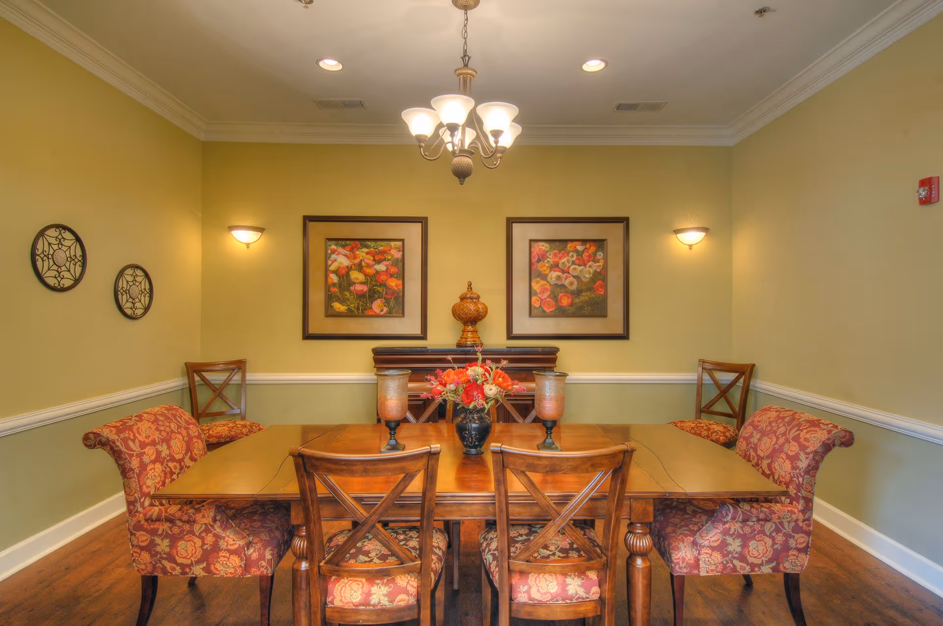 A dining room with a wooden table surrounded by six chairs, four with floral upholstery and two wooden chairs with cross backs. The table has a floral centerpiece and two candle holders. The walls are painted light green with white trim, decorated with two framed floral paintings and two decorative wall hangings. A chandelier with five lights hangs from the ceiling.