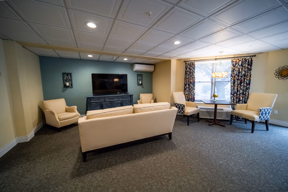 A cozy living room area with beige upholstered sofa and armchairs arranged around a flat-screen TV mounted on a blue accent wall. There is a small table with three chairs near a window with patterned curtains, and the room has recessed lighting and a carpeted floor.