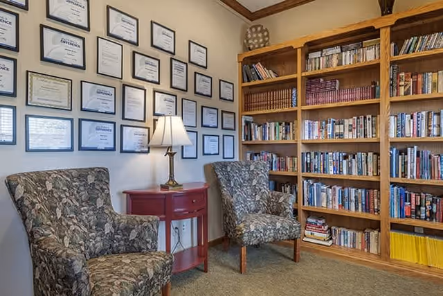 A cozy reading area with two floral patterned armchairs facing each other, a small red wooden side table with a lamp between them, a wall covered with framed certificates, and a large wooden bookshelf filled with books.