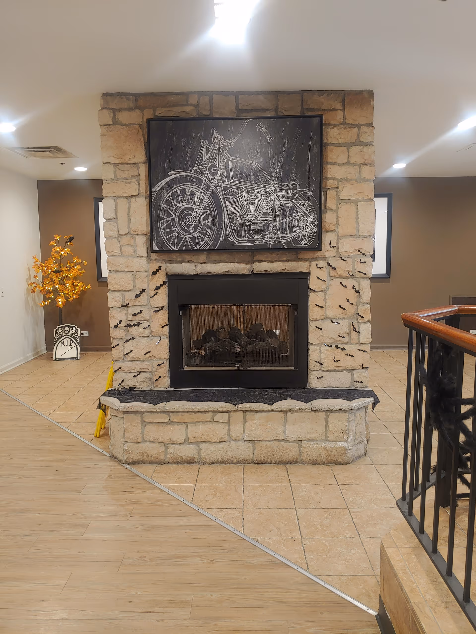 Interior view of a room with a stone fireplace decorated with small black bat cutouts. Above the fireplace is a framed chalk-style drawing of a motorcycle. To the left, there is a small tree with orange lights and a tombstone decoration marked RIP, suggesting Halloween decor. The floor is a combination of wood and tile, and there is a black railing on the right side.