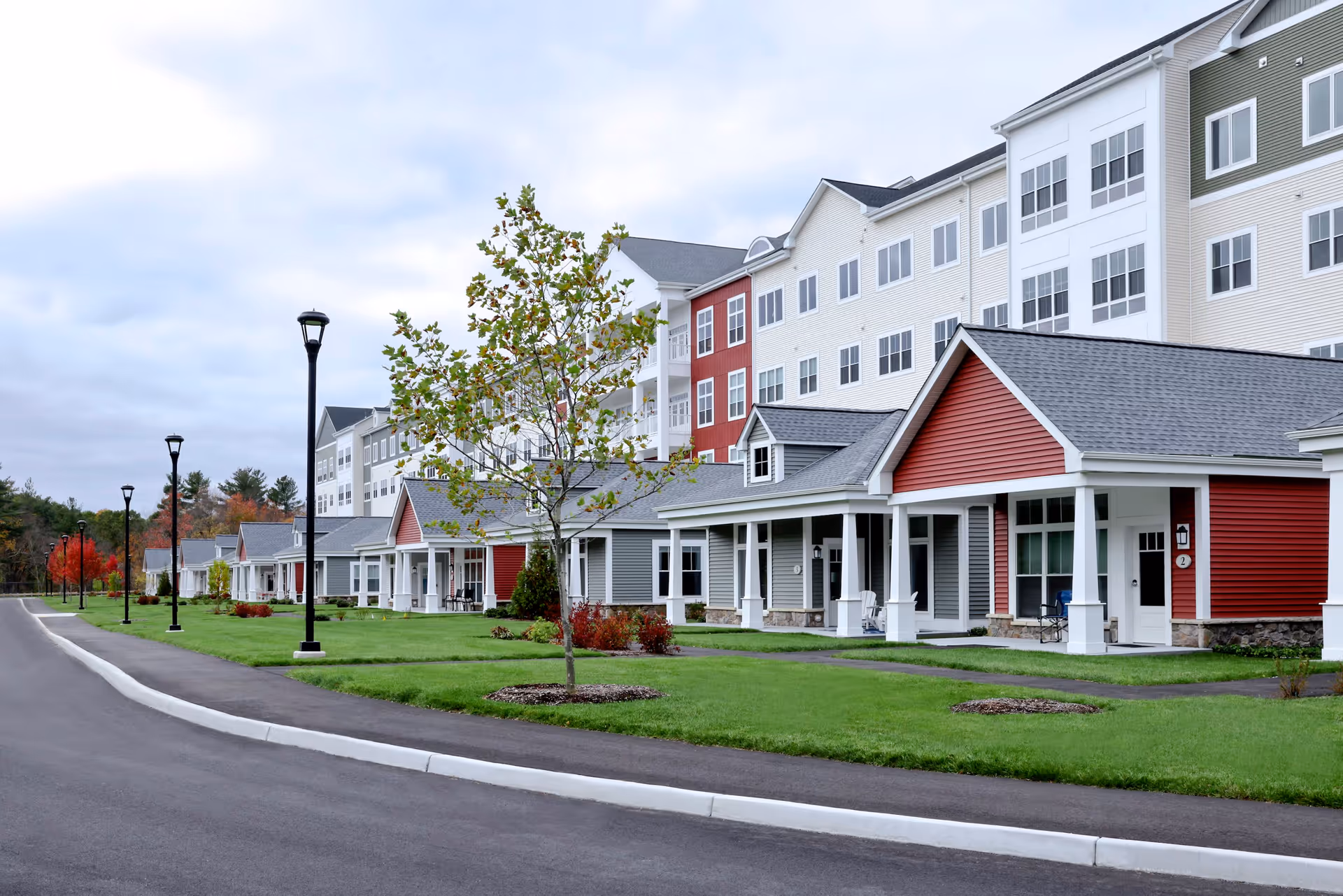 Exterior view of The Willows Retirement Community showing a row of single-story cottages with porches and a multi-story building behind them. The buildings have a mix of red, gray, white, and green siding with multiple windows. There is a paved road and sidewalk with street lamps and green lawns with small trees and shrubs.