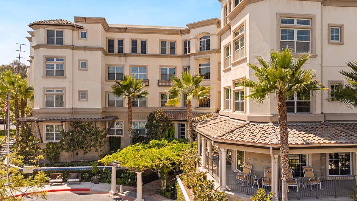 Exterior view of a multi-story senior living facility building with beige walls and numerous windows, surrounded by palm trees and greenery. There is a covered patio area with tables and chairs on the right side, and benches and landscaped pathways in the courtyard area.