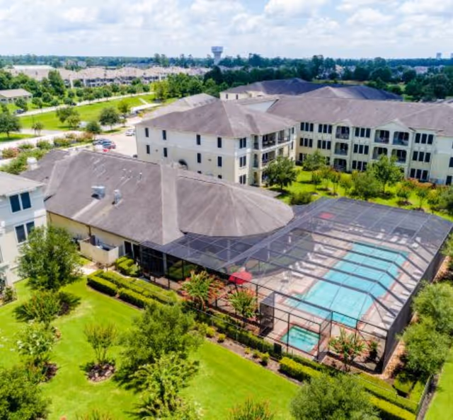 Aerial view of Arella Forest at Woodland an Active Adult Apartment complex featuring multiple beige buildings with dark roofs, surrounded by green lawns and trees. The image prominently shows a screened-in outdoor swimming pool area with lounge chairs and a hot tub.