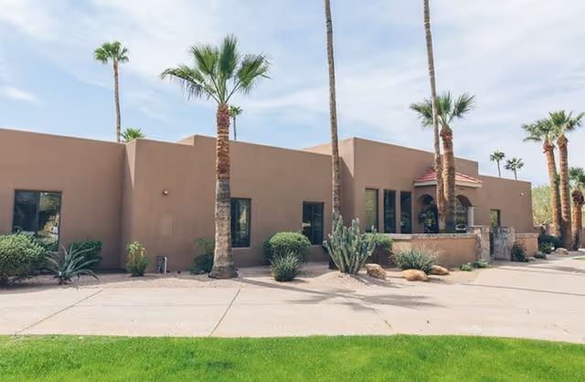 Exterior view of a single-story building with a flat roof, beige stucco walls, and several windows. The building is surrounded by desert landscaping including palm trees, cacti, and shrubs. There is a paved walkway and a green lawn in the foreground under a partly cloudy sky.