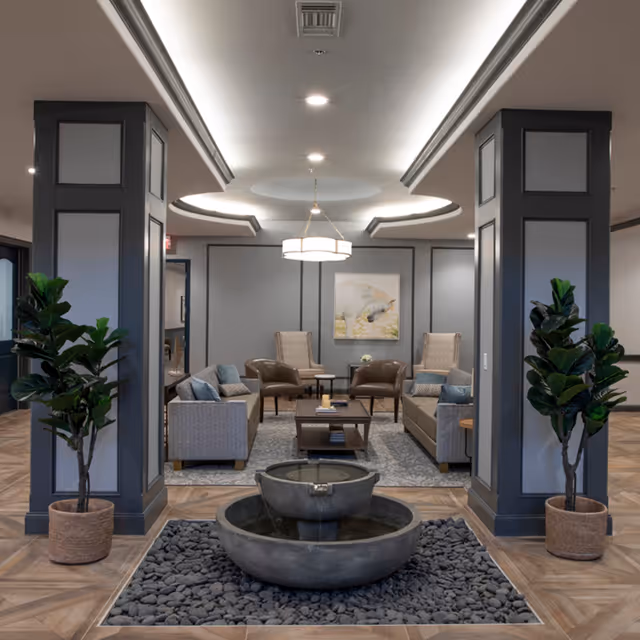 A modern and elegant living room area in a senior living facility featuring two gray sofas, two brown armchairs, two beige armchairs, a central coffee table, and a decorative water fountain surrounded by black pebbles in the foreground. The room has gray paneled walls, a circular ceiling light fixture, and two large potted plants flanking the fountain.