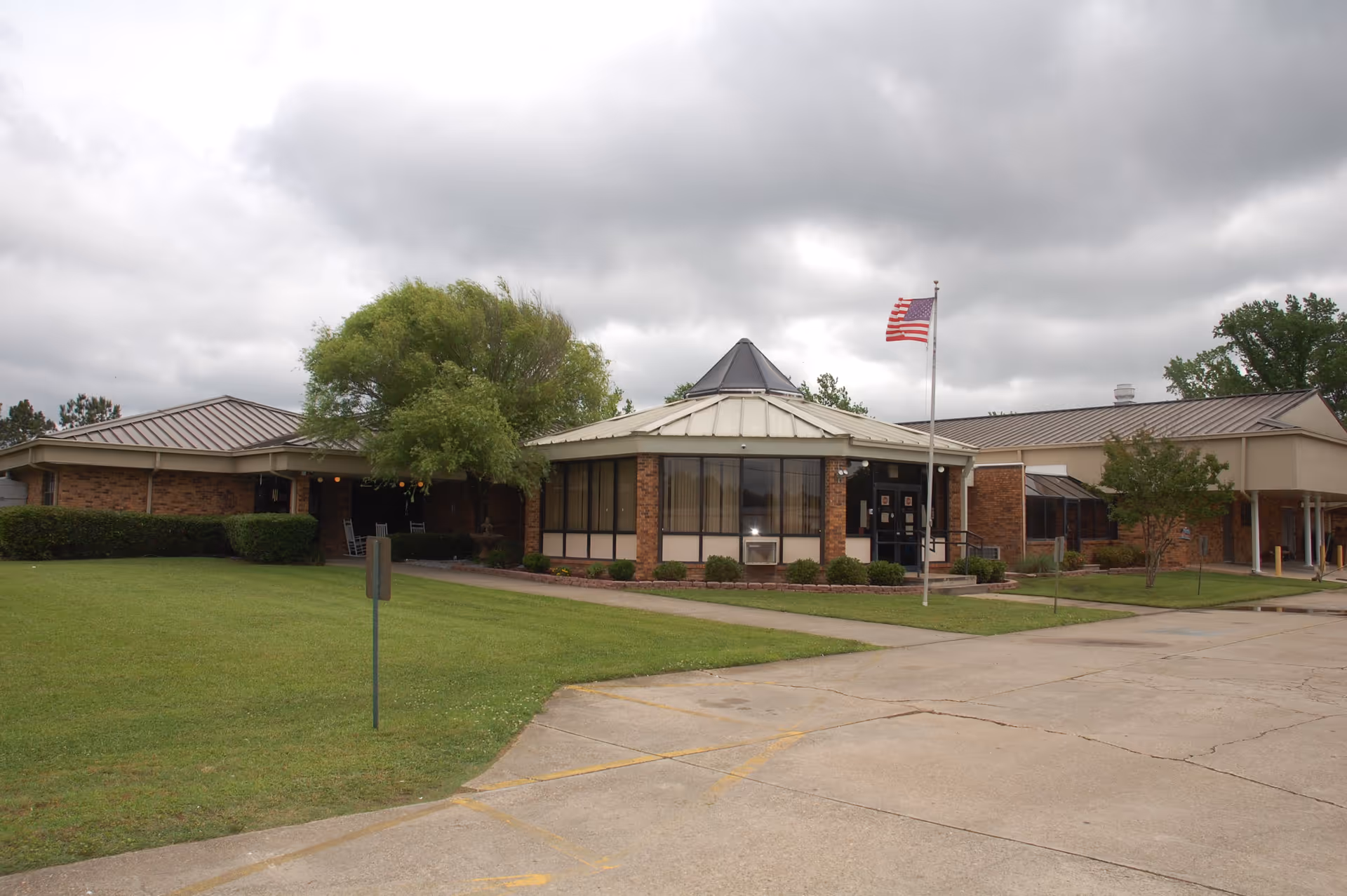 Front exterior of a single-story brick senior living facility with a central glass entry, flagpole flying an American flag, and a lawn under a cloudy sky.