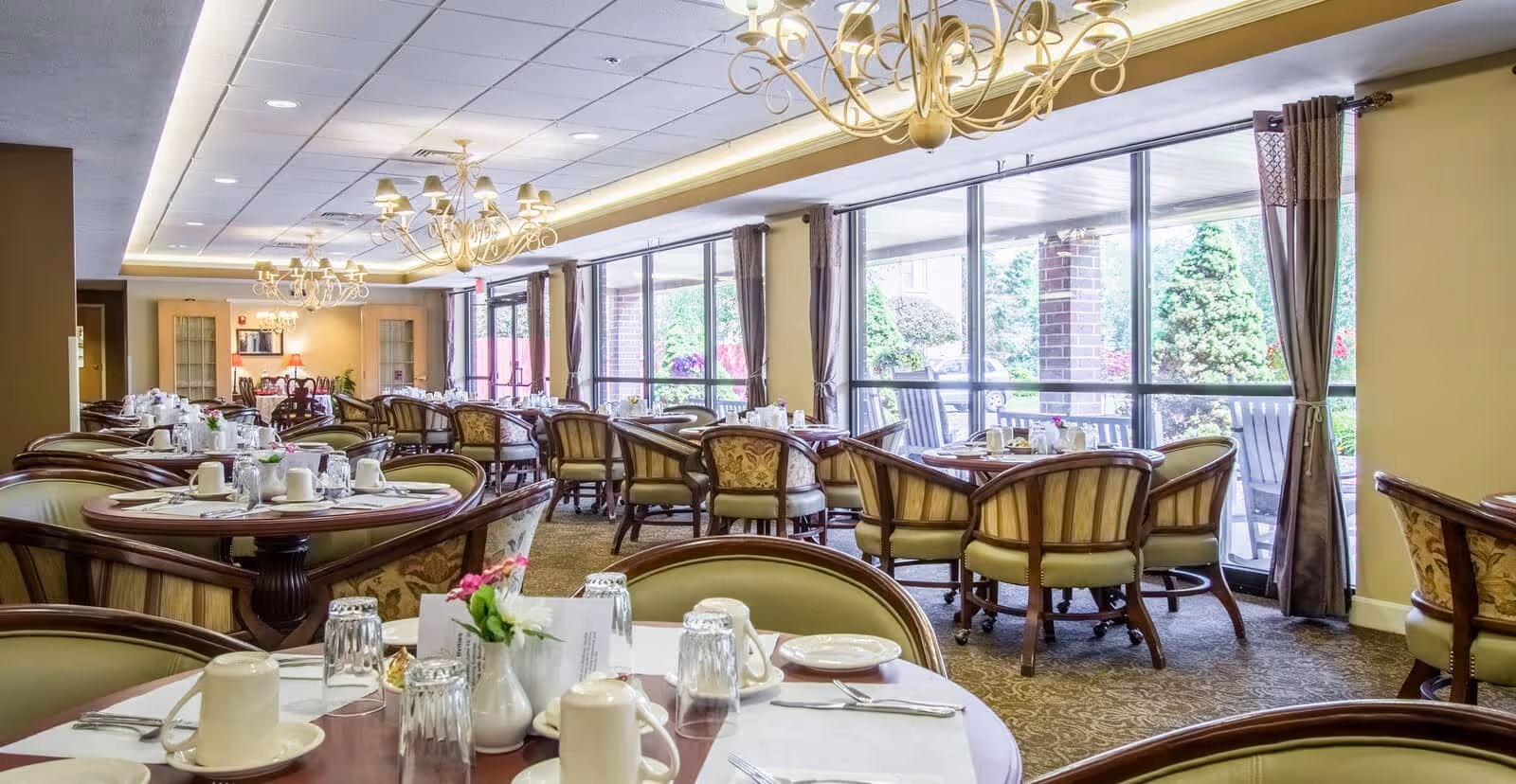 A bright and elegant dining room in a senior living community with multiple round tables set with white cups, glasses, plates, and silverware. The room features large windows with curtains allowing natural light to illuminate the space, ornate chandeliers hanging from the ceiling, and upholstered chairs around each table. Outside the windows, greenery and garden elements are visible.