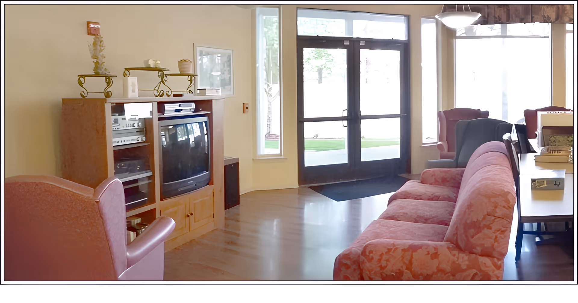 A cozy living room area with a floral-patterned pink sofa, two armchairs, a wooden entertainment center with a TV and stereo equipment, and large glass doors leading outside. The room has wooden flooring and is well-lit with natural light from the windows.