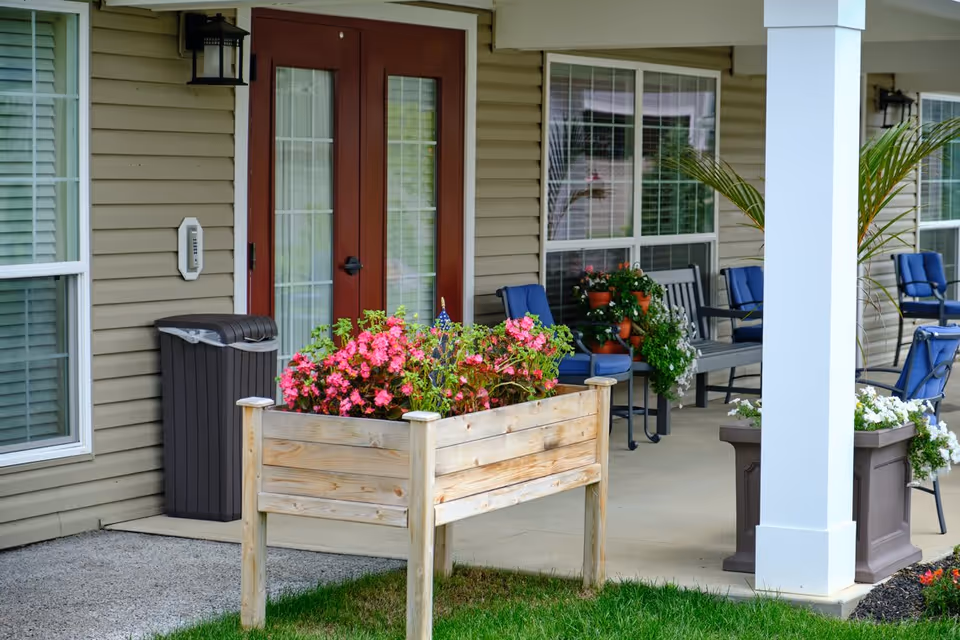 Outdoor patio area of a senior living facility with a raised wooden planter box filled with pink flowers in the foreground. The patio has several blue cushioned chairs, a gray bench, potted plants, and a beige building exterior with red double doors and large windows.
