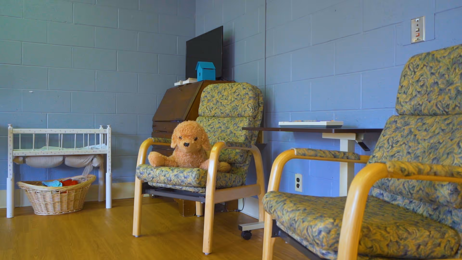A room with two patterned armchairs, a small table between them, a teddy bear sitting on one chair, a basket with folded clothes, and a small white crib against a blue painted brick wall.