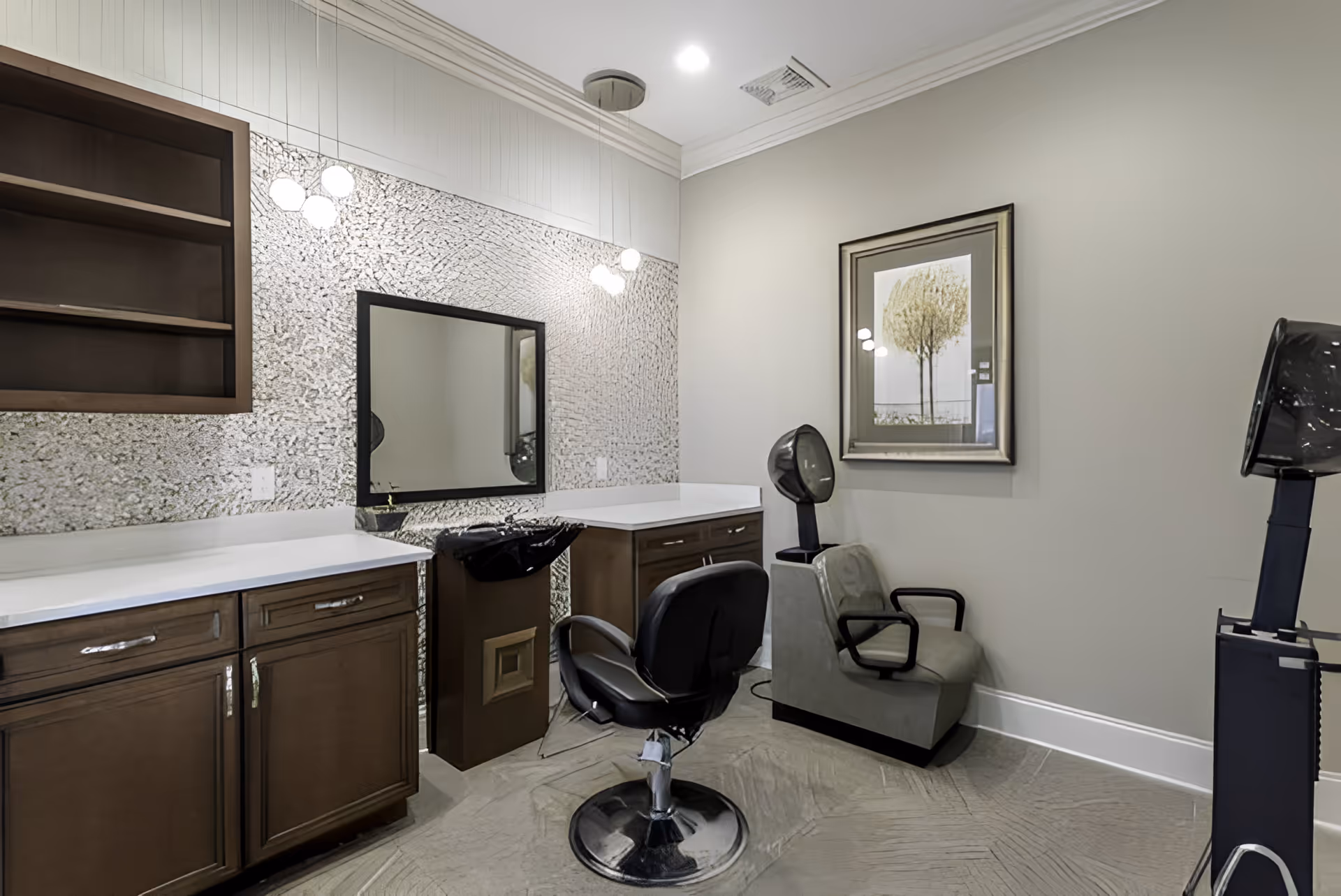 Interior view of a salon area in an assisted living facility featuring a black salon chair in front of a mirror and countertop with wooden cabinets. To the right, there is a gray hair dryer chair with a hooded hair dryer. A framed picture of a tree hangs on the wall above the hair dryer chair.