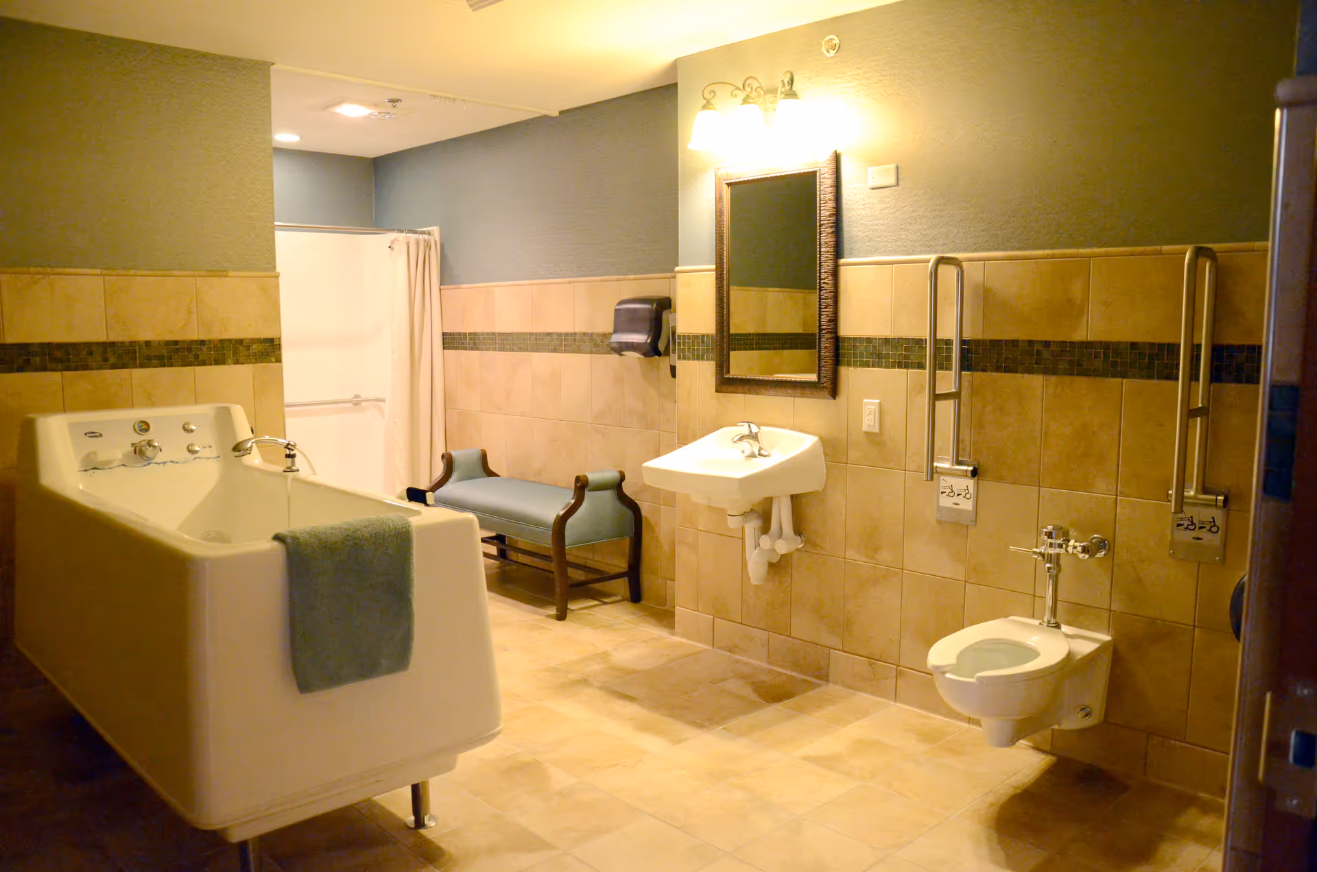 A spacious bathroom with beige tiled walls and floor, featuring a large white bathtub with a green towel draped over the side, a wall-mounted sink with a mirror above it, a small blue bench, and a wall-mounted toilet with support bars on either side.
