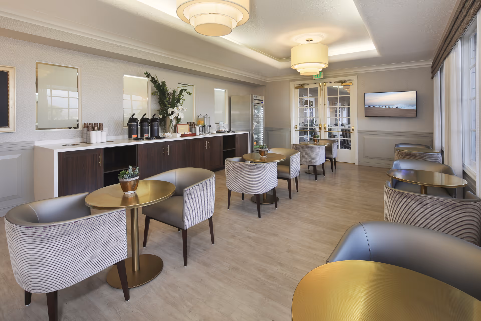 A bright and modern dining area with several small round tables and upholstered chairs arranged neatly. Along one wall, there is a long counter with coffee dispensers, cups, and a variety of beverages. The room features light-colored walls, large windows with curtains, and two large ceiling light fixtures. A flat-screen TV is mounted on the far wall near double glass doors.