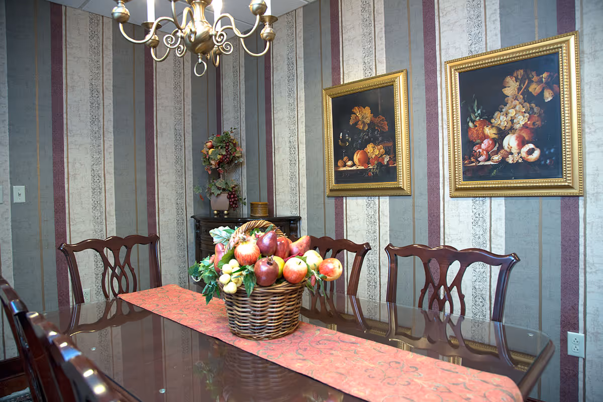 A dining room with a polished wooden table and six matching chairs. A basket filled with artificial fruits is placed on a red table runner in the center of the table. The walls have vertical striped wallpaper in shades of gray, beige, and burgundy. Two framed paintings of fruit still lifes hang on the wall, and a chandelier with gold accents hangs above the table.