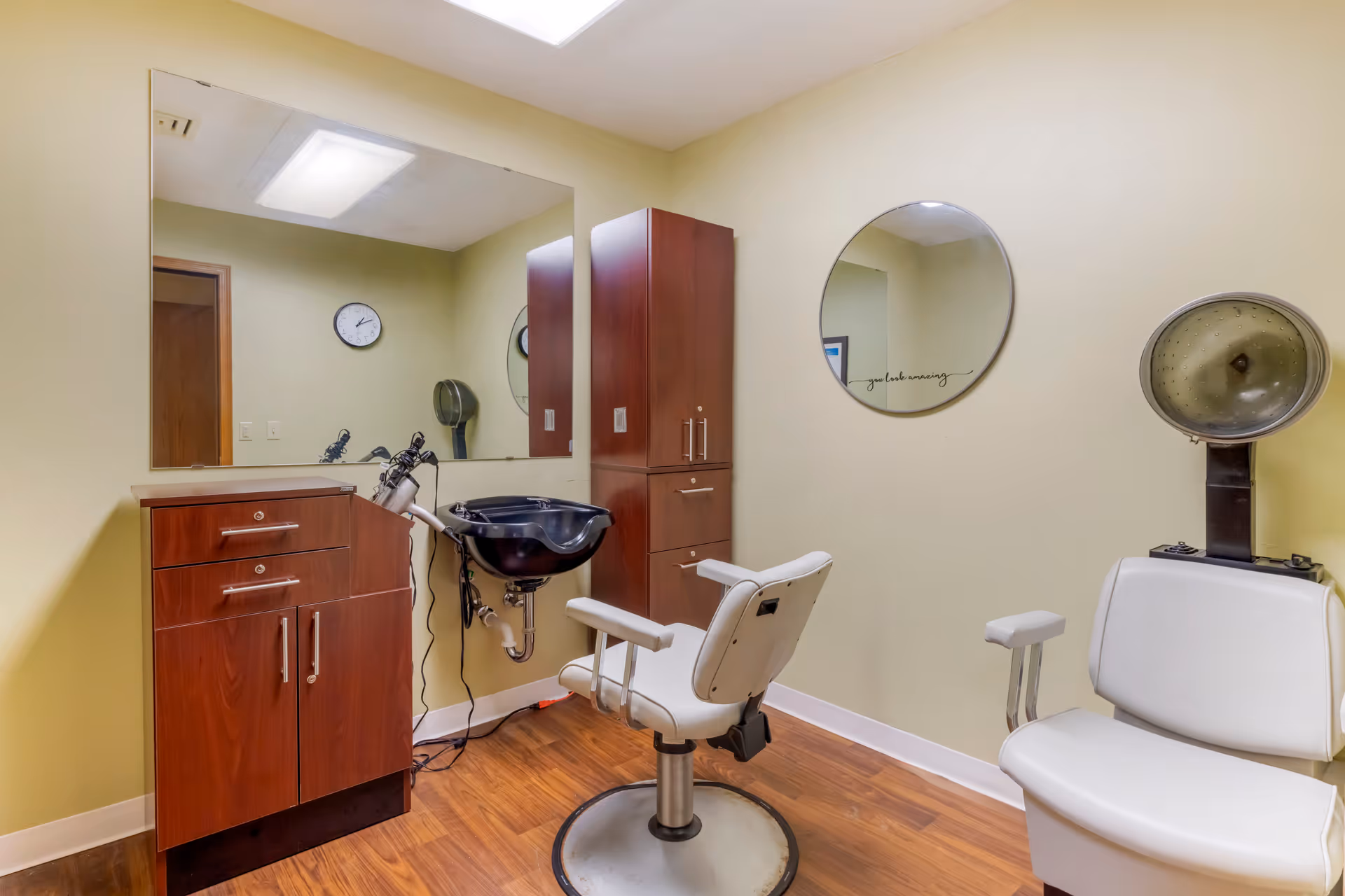 Interior of a hair salon room with a white salon chair in front of a black sink, wooden cabinets, a large rectangular mirror on the wall, a round mirror, and a white hair dryer chair with a hood dryer attached. The room has light yellow walls and wooden flooring.