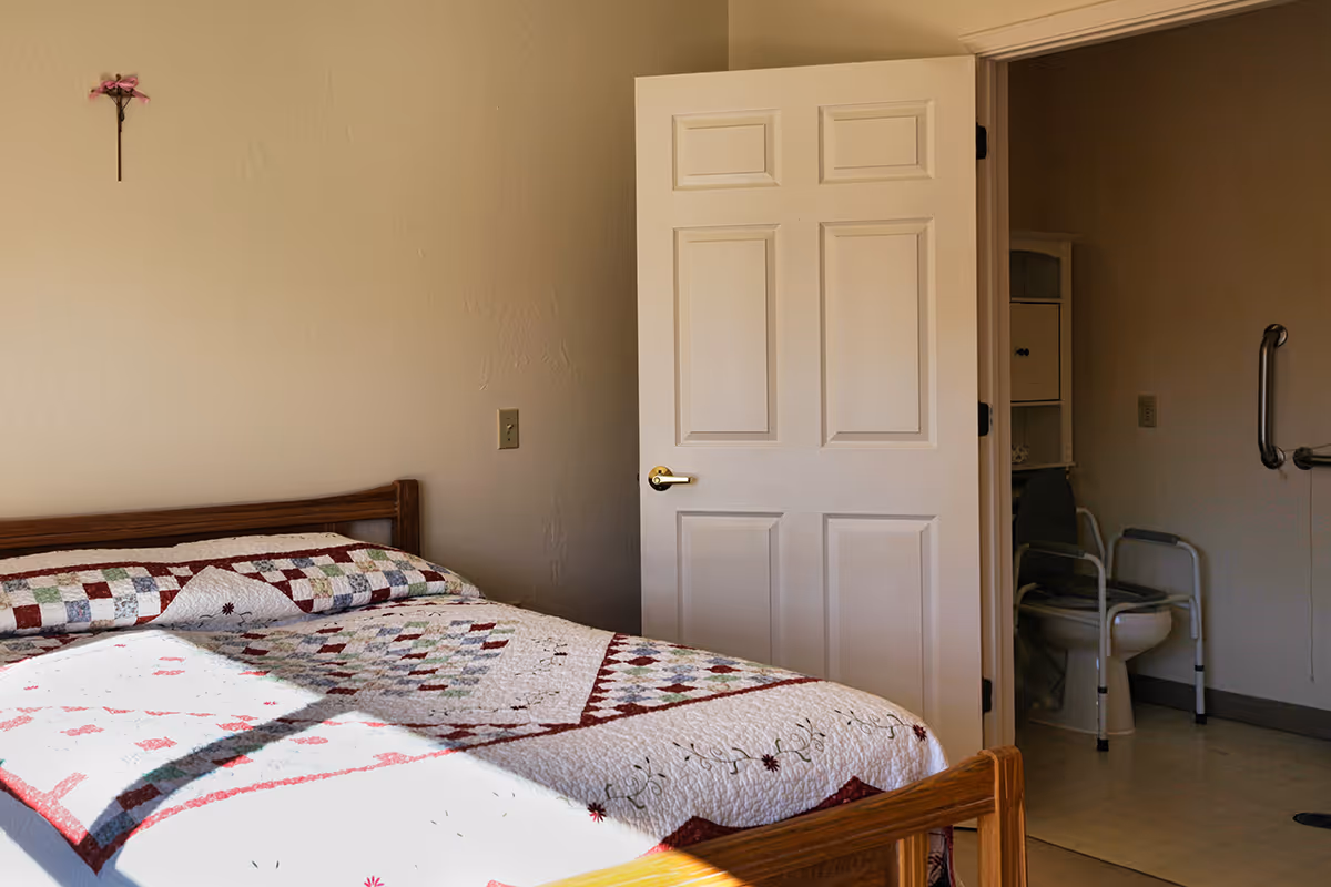 A bedroom with a wooden bed frame and a colorful patchwork quilt. The door to an adjacent bathroom is open, showing a toilet with a safety frame and grab bars on the wall. The room has beige walls and a small decorative flower hanging on the wall above the bed.