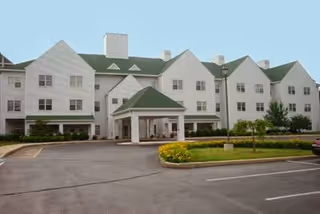 Front exterior of a multi-story white assisted living building with green roofs, a covered porte-cochère entrance, parking area, and landscaped flower beds.
