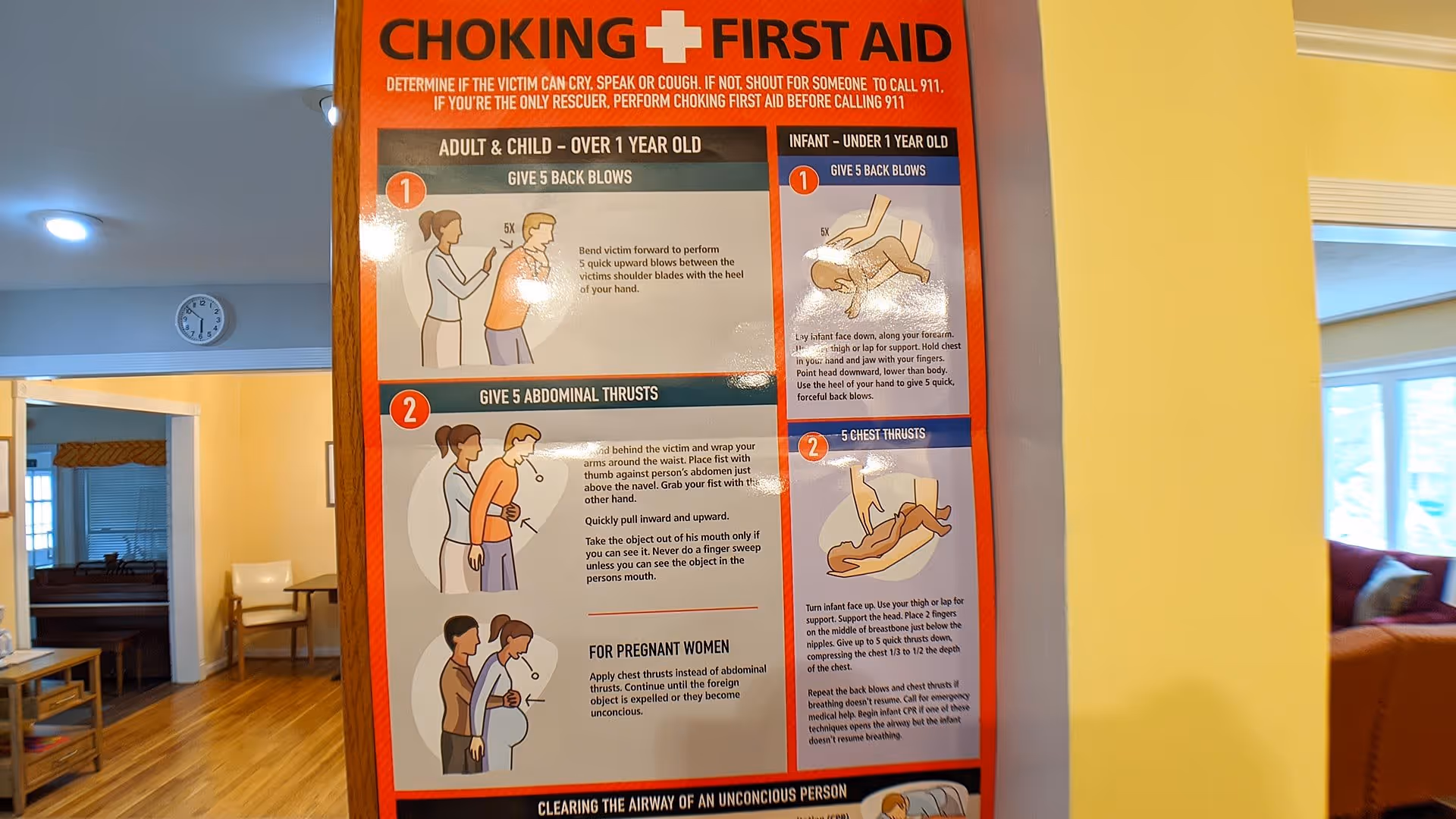 A large 'Choking + First Aid' instructional poster mounted on a wooden pillar in a senior living facility common area with seating and windows in the background.